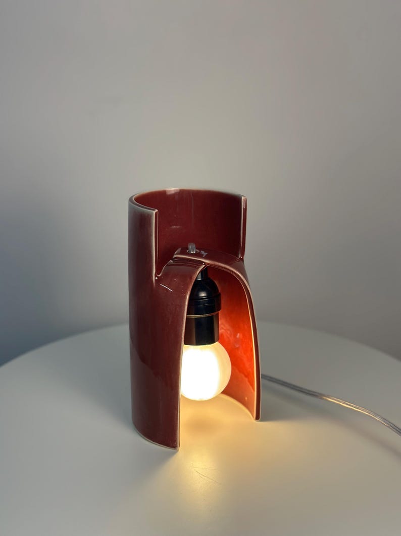 May include: A cylindrical ceramic table lamp with a glossy dark red finish. The lamp features a cutout arch design that exposes a black socket holding a glowing light bulb. A thin, light-colored electrical cord extends from the base of the lamp.