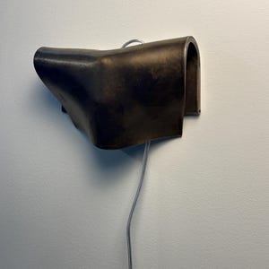 May include: A dark brown, curved wall-mounted lamp with a smooth, matte finish. The fixture features a folded, sculptural design that extends outwards from the wall, with a thin, silver-toned electrical cord hanging vertically from the bottom.