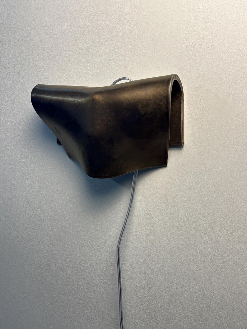 May include: A dark brown, curved wall-mounted lamp with a smooth, matte finish. The fixture features a folded, sculptural design that extends outwards from the wall, with a thin, silver-toned electrical cord hanging vertically from the bottom.