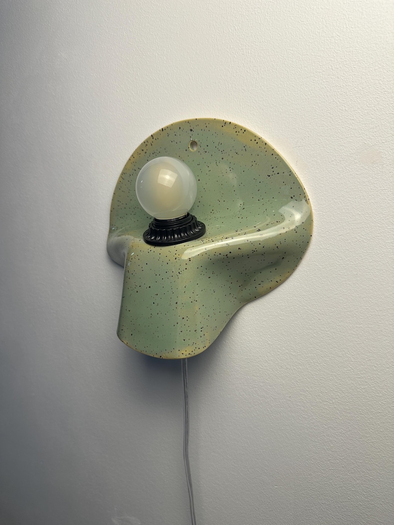 Puede incluir: Un aplique de pared con una base de cer&aacute;mica de color verde claro moteado, con una forma curva y un acabado brillante. Una bombilla redonda de cristal blanco esmerilado se asienta en un casquillo negro centrado, con un cable fino.