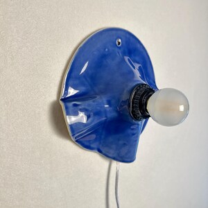May include: A blue ceramic wall sconce with a bulb. The sconce has a unique, organic shape and a glossy finish. The bulb is round and emits a soft light. A gray wire extends from the sconce.