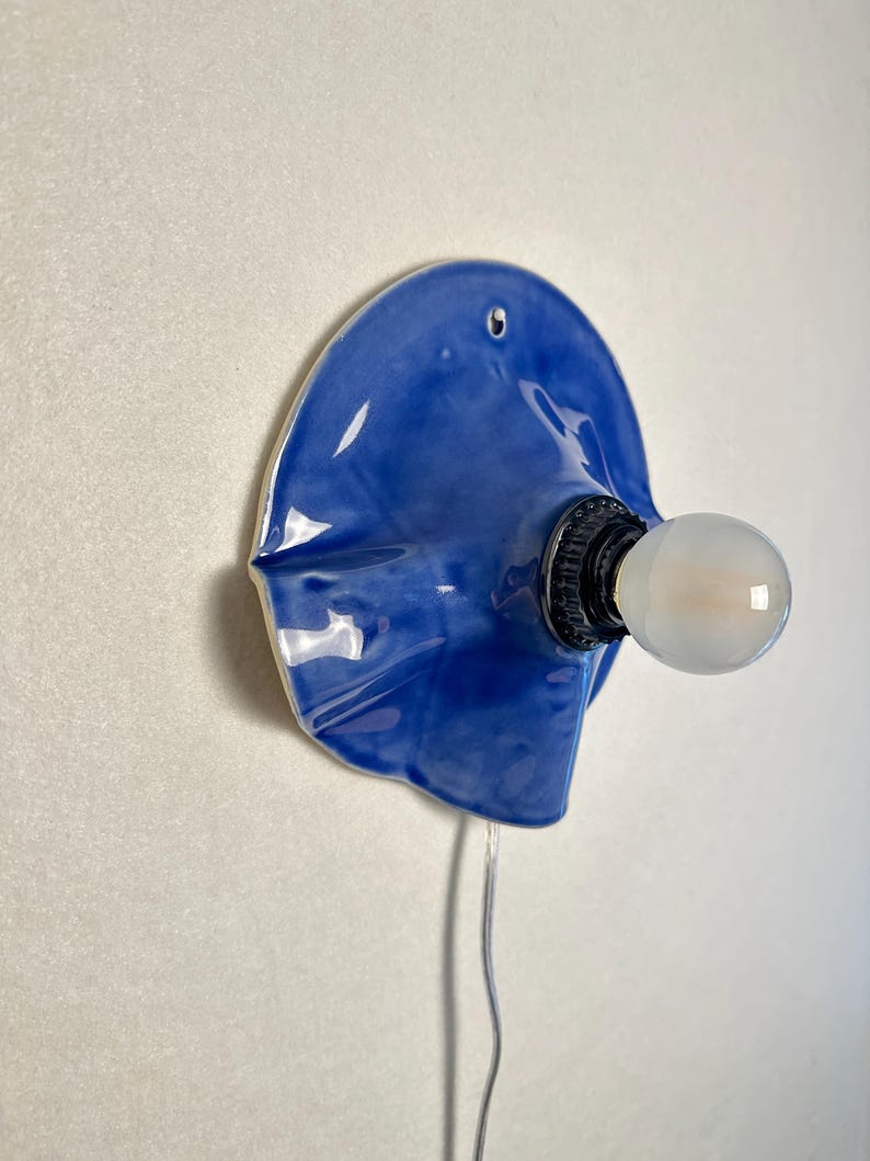 May include: A blue ceramic wall sconce with a bulb. The sconce has a unique, organic shape and a glossy finish. The bulb is round and emits a soft light. A gray wire extends from the sconce.