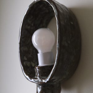 May include: A handcrafted, dark brown ceramic wall sconce with a white light bulb. The sconce has an oval shape with a textured surface and a glossy finish. The light bulb is visible inside the sconce.
