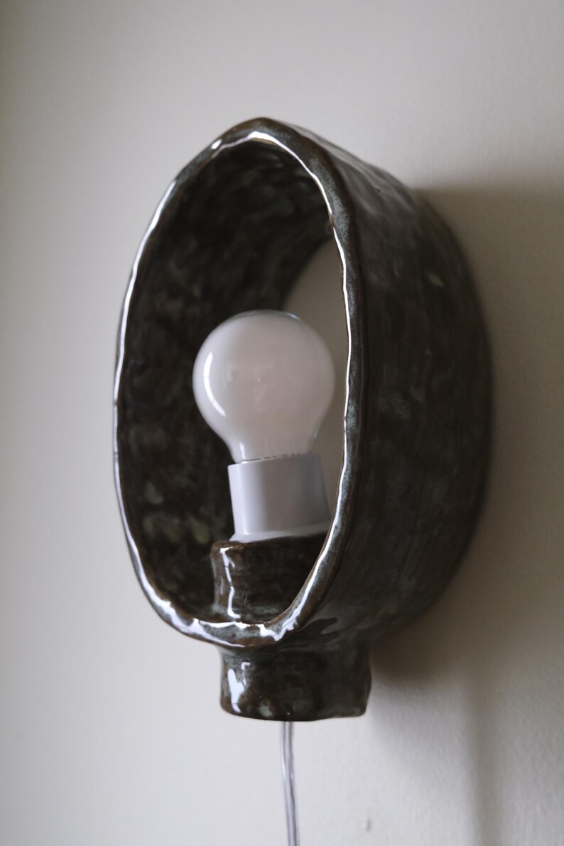 May include: A handcrafted, dark brown ceramic wall sconce with a white light bulb. The sconce has an oval shape with a textured surface and a glossy finish. The light bulb is visible inside the sconce.