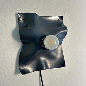May include: A wall-mounted lamp featuring a dark gray, wavy, square-shaped metal backplate with a central black socket holding a round, frosted white light bulb. A thin black power cord extends downward from the base of the fixture.
