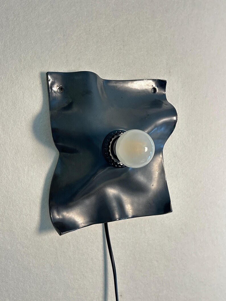 May include: A wall-mounted lamp featuring a dark gray, wavy, square-shaped metal backplate with a central black socket holding a round, frosted white light bulb. A thin black power cord extends downward from the base of the fixture.