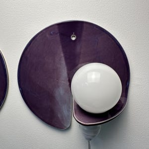 K&ouml;nnte beinhalten: Eine Wandleuchte mit einer kreisf&ouml;rmigen, tiefvioletten Keramik-R&uuml;ckplatte mit gl&auml;nzender Oberfl&auml;che. Eine einzelne, kugelf&ouml;rmige wei&szlig;e Gl&uuml;hbirne ist an der unteren rechten Seite der Platte angebracht und mit einer wei&szlig;en Fassung und Kabel verbunden.