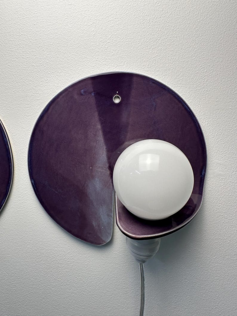 K&ouml;nnte beinhalten: Eine Wandleuchte mit einer kreisf&ouml;rmigen, tiefvioletten Keramik-R&uuml;ckplatte mit gl&auml;nzender Oberfl&auml;che. Eine einzelne, kugelf&ouml;rmige wei&szlig;e Gl&uuml;hbirne ist an der unteren rechten Seite der Platte angebracht und mit einer wei&szlig;en Fassung und Kabel verbunden.