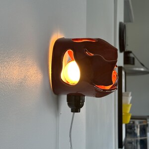 May include: A dark brown wall sconce with an abstract design. The sconce emits a warm, orange glow, with a visible light bulb. The fixture is mounted on a white wall, with a black electrical cord and a plug.
