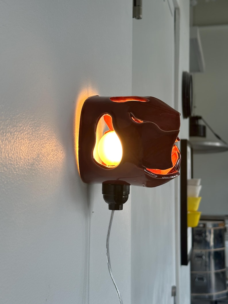 May include: A dark brown wall sconce with an abstract design. The sconce emits a warm, orange glow, with a visible light bulb. The fixture is mounted on a white wall, with a black electrical cord and a plug.