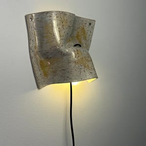 May include: A wall-mounted light fixture with a curved, irregular, and folded ceramic shade. The surface features a speckled pattern in shades of beige, gray, and yellow. A black electrical cord extends downward from the base of the illuminated lamp.