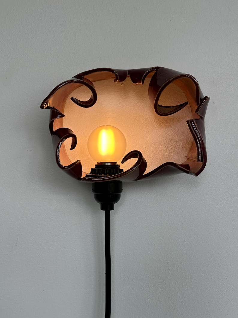 May include: A unique, handcrafted wall sconce featuring a dark brown, petal-shaped shade. The shade surrounds a warm, glowing light bulb. The sconce has a black base and a black cord. The overall design is artistic and decorative.