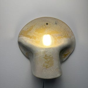 May include: A wall-mounted ceramic sconce with a rounded, organic shape and a textured, mottled cream and yellow glaze. A single glowing, spherical light bulb is recessed into the center of the fixture, which features a small hole at the top.