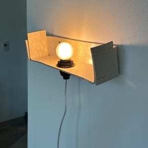 May include: A modern wall-mounted light fixture with a speckled, light-colored shelf. A round, illuminated bulb sits on a dark base, casting a warm glow. The fixture is attached to a wall with a visible power cord.
