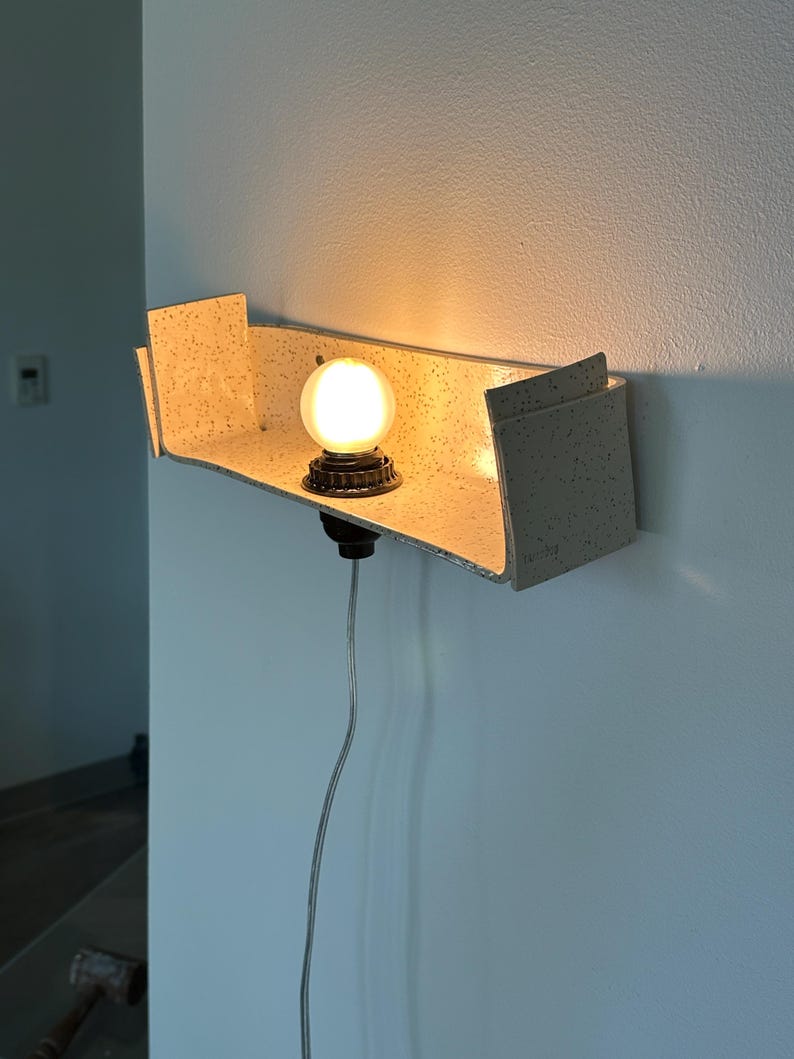May include: A modern wall-mounted light fixture with a speckled, light-colored shelf. A round, illuminated bulb sits on a dark base, casting a warm glow. The fixture is attached to a wall with a visible power cord.