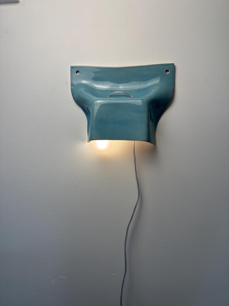 K&ouml;nnte beinhalten: Eine Wandleuchte mit einem gl&auml;nzenden, hellblauen Keramikschirm in geschwungener, geformter Optik. Die Leuchte verf&uuml;gt &uuml;ber einen kleinen Metallring an der Oberseite, zwei Befestigungsl&ouml;cher und eine leuchtende Gl&uuml;hbirne mit Kabel.