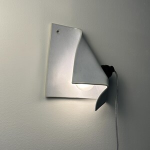 May include: A white ceramic wall sconce with a folded, sheet-like design. The fixture features a single light bulb tucked behind the curved edge, a small circular mounting hole at the top, and a black socket connected to a thin white power cord.