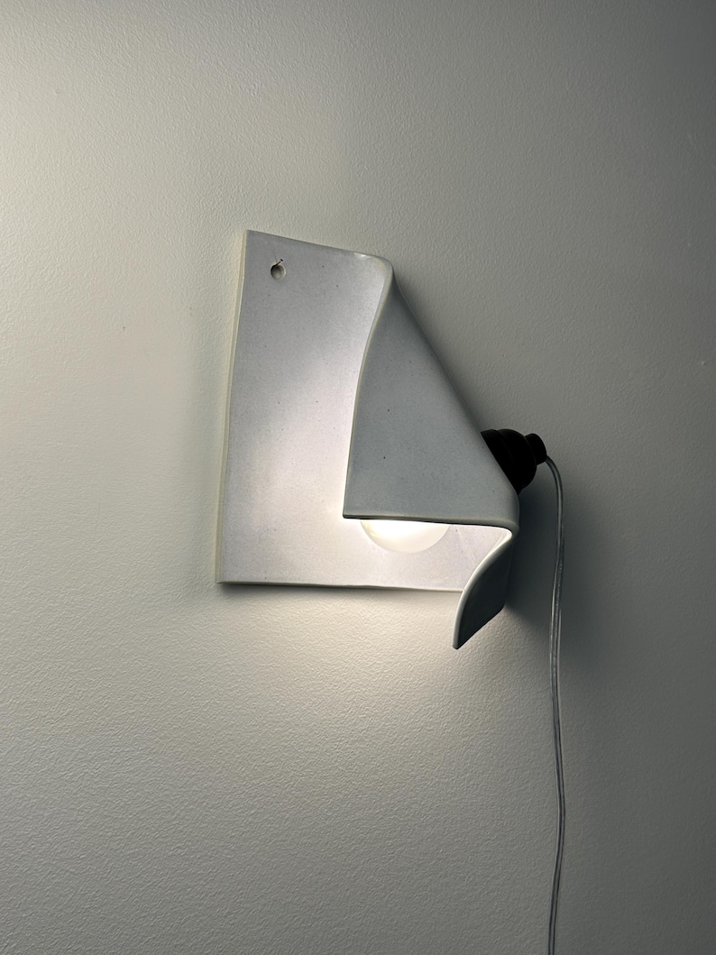 May include: A white ceramic wall sconce with a folded, sheet-like design. The fixture features a single light bulb tucked behind the curved edge, a small circular mounting hole at the top, and a black socket connected to a thin white power cord.