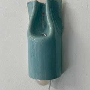 May include: A wall-mounted light fixture with a glossy, light blue ceramic shade featuring a fluid, organic, and undulating shape. The fixture includes a small, round, frosted glass bulb protruding from the bottom and a clear power cord.