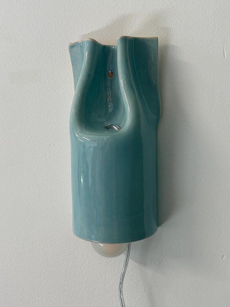 May include: A wall-mounted light fixture with a glossy, light blue ceramic shade featuring a fluid, organic, and undulating shape. The fixture includes a small, round, frosted glass bulb protruding from the bottom and a clear power cord.