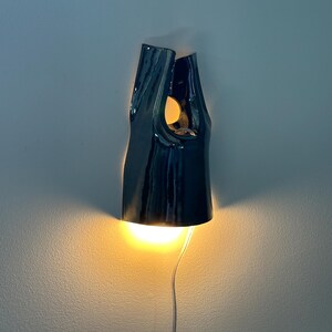 May include: A dark, glossy ceramic wall sconce with an irregular, elongated shape. The fixture features an open top and a cutout section, emitting a warm, bright yellow light from both the bottom opening and the upper cutout, with a thin power cord.