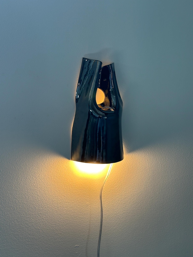 May include: A dark, glossy ceramic wall sconce with an irregular, elongated shape. The fixture features an open top and a cutout section, emitting a warm, bright yellow light from both the bottom opening and the upper cutout, with a thin power cord.