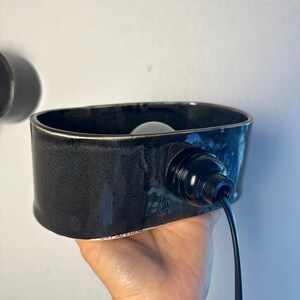 May include: A black ceramic wall sconce with a rectangular shape. The sconce has a black cord and a light bulb inside. The sconce is held in a hand.