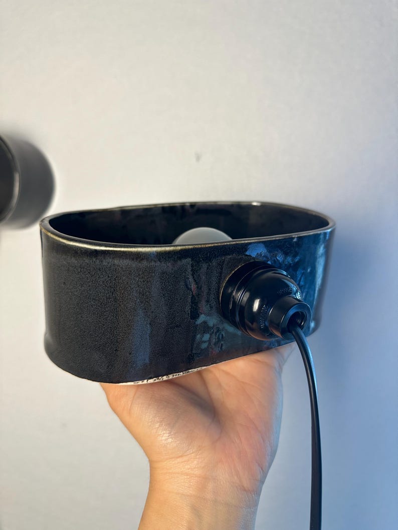May include: A black ceramic wall sconce with a rectangular shape. The sconce has a black cord and a light bulb inside. The sconce is held in a hand.