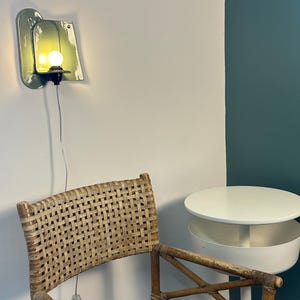 May include: A wall-mounted light fixture with a glossy, sage green ceramic backplate and a single glowing bulb. The lamp features a black socket and a thin, transparent power cord with an inline switch, mounted on a plain white wall.