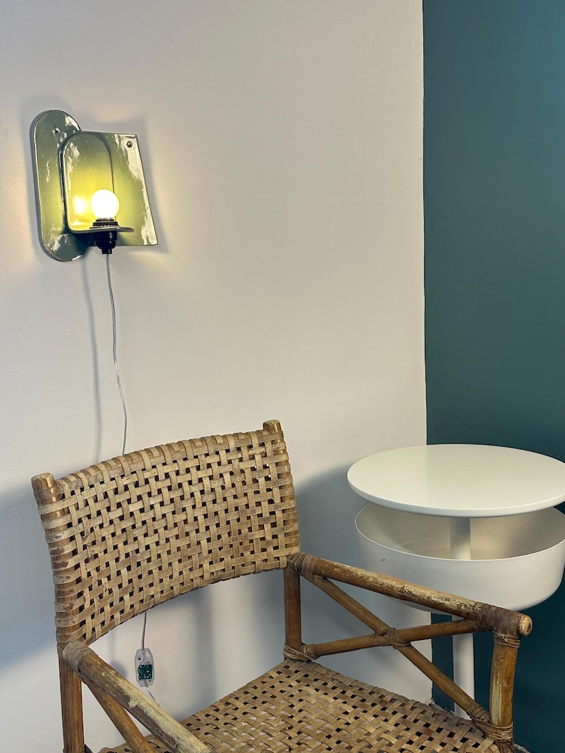May include: A wall-mounted light fixture with a glossy, sage green ceramic backplate and a single glowing bulb. The lamp features a black socket and a thin, transparent power cord with an inline switch, mounted on a plain white wall.