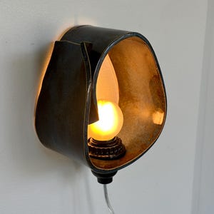 May include: A unique wall sconce with a dark, textured exterior and a warm, golden interior. The sconce houses a small, round light bulb, casting a soft glow. The fixture is mounted on a white wall, with a cord extending downwards.