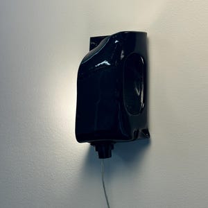 May include: A glossy black wall-mounted light fixture with a curved, sculptural form. The fixture features a smooth, reflective surface and a central opening, with a small black component at the base connected to a thin, downward-extending power cord.