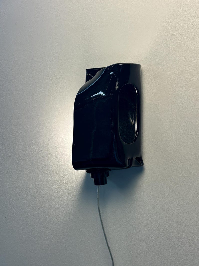 May include: A glossy black wall-mounted light fixture with a curved, sculptural form. The fixture features a smooth, reflective surface and a central opening, with a small black component at the base connected to a thin, downward-extending power cord.