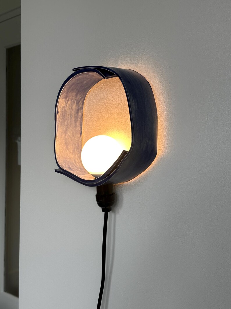 Puede incluir: Una l&aacute;mpara de pared moderna con una pantalla curva azul oscuro. La pantalla rodea una bombilla redonda y blanca. Un cable negro se extiende hacia abajo desde la l&aacute;mpara.