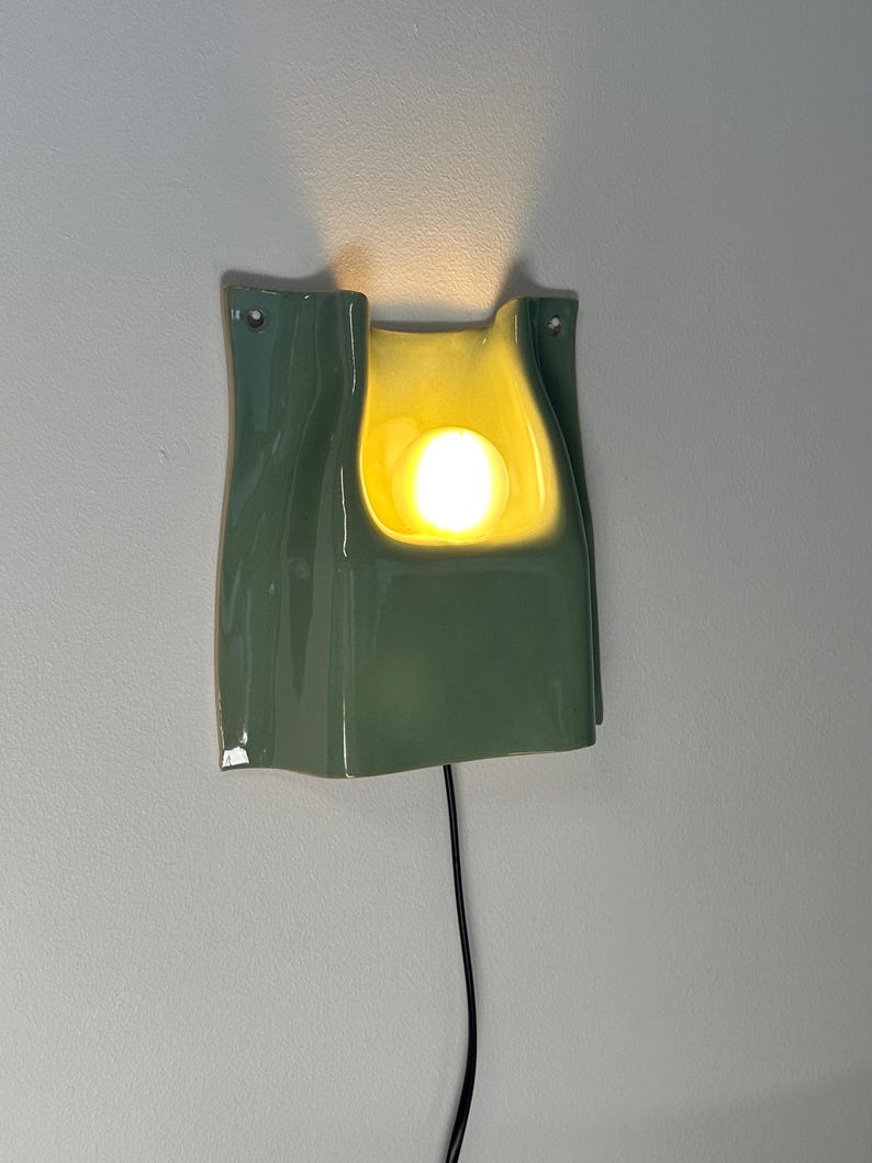 May include: A sage green ceramic wall sconce with a glossy finish and a curved, folded silhouette. The fixture features a central cutout housing a glowing, warm-toned light bulb. A thin black power cord extends downwards from the base of the light.