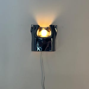 May include: A wall-mounted lamp featuring a glossy, dark navy blue ceramic housing with an irregular, organic shape. The fixture emits a warm, glowing light from a central circular bulb, and includes a thin, clear power cord extending downward.