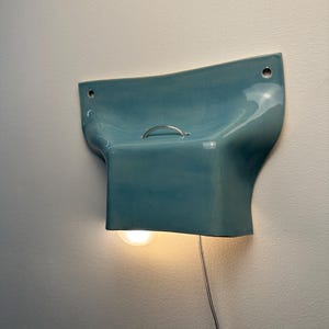 K&ouml;nnte beinhalten: Eine Wandleuchte mit einem gl&auml;nzenden, hellblauen Keramikschirm in geschwungenem, ausgestelltem Design. Die Leuchte verf&uuml;gt &uuml;ber einen kleinen silbernen Metallgriff an der Oberseite, zwei Befestigungsl&ouml;cher und eine leuchtende Gl&uuml;hbirne.