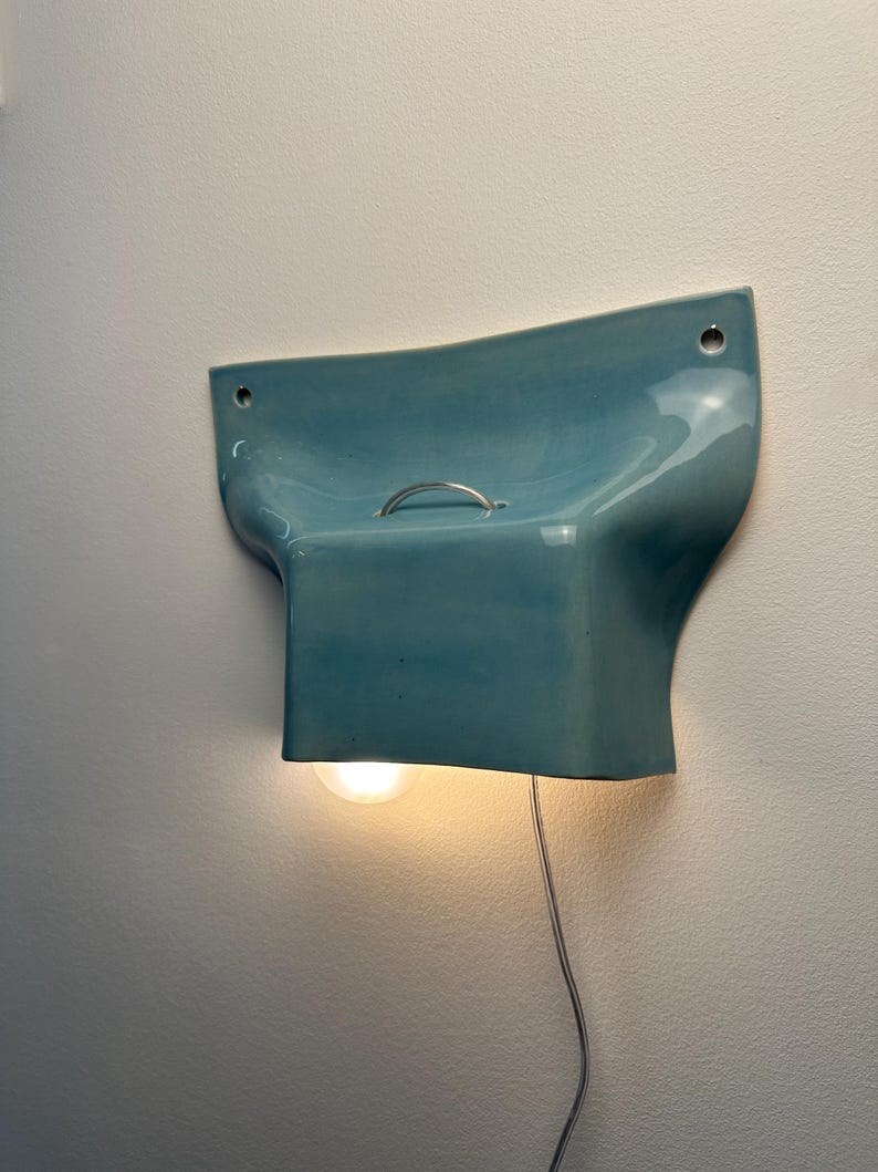 K&ouml;nnte beinhalten: Eine Wandleuchte mit einem gl&auml;nzenden, hellblauen Keramikschirm in geschwungenem, ausgestelltem Design. Die Leuchte verf&uuml;gt &uuml;ber einen kleinen silbernen Metallgriff an der Oberseite, zwei Befestigungsl&ouml;cher und eine leuchtende Gl&uuml;hbirne.