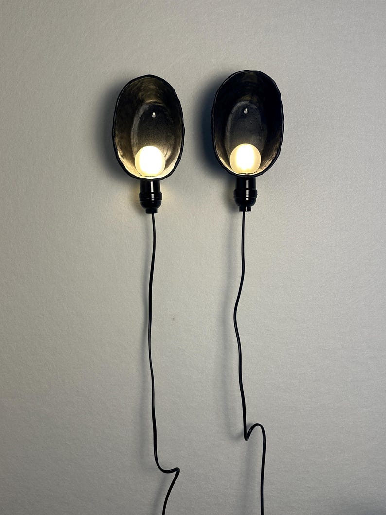 May include: Two wall-mounted lamps featuring oval-shaped, dark-colored, textured shades. Each lamp contains a single glowing, spherical light bulb positioned within the shade, connected to a black cord that hangs vertically downward from the base.