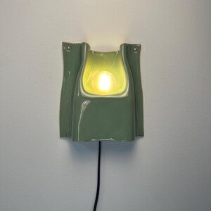 May include: A sage green ceramic wall sconce with a glossy finish and a curved, folded silhouette. The fixture features a central cutout housing a glowing, warm-toned light bulb. A thin black power cord extends downwards from the base of the lamp.