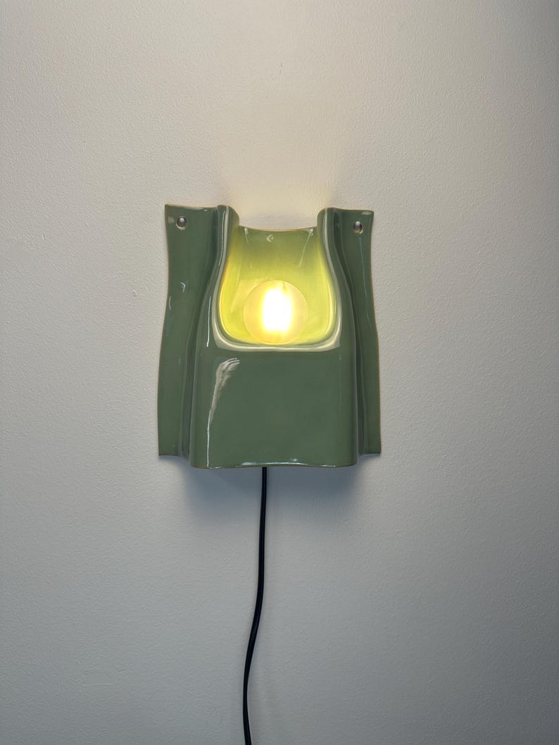 May include: A sage green ceramic wall sconce with a glossy finish and a curved, folded silhouette. The fixture features a central cutout housing a glowing, warm-toned light bulb. A thin black power cord extends downwards from the base of the lamp.