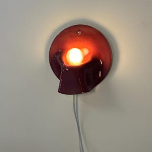 May include: A circular wall lamp with a deep burgundy, glossy ceramic finish. The lamp features a central, protruding hood that houses a single glowing light bulb. A small hole is visible at the top for mounting, and a thin white power cord hangs below.