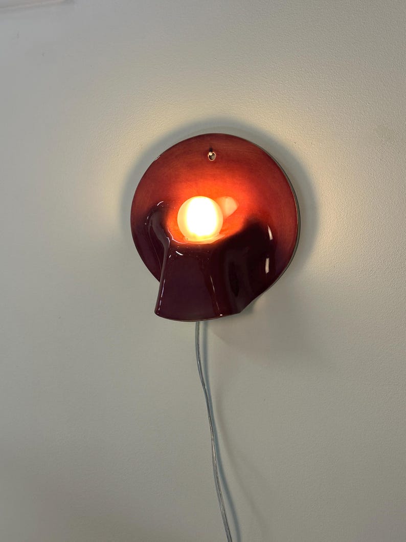 May include: A circular wall lamp with a deep burgundy, glossy ceramic finish. The lamp features a central, protruding hood that houses a single glowing light bulb. A small hole is visible at the top for mounting, and a thin white power cord hangs below.