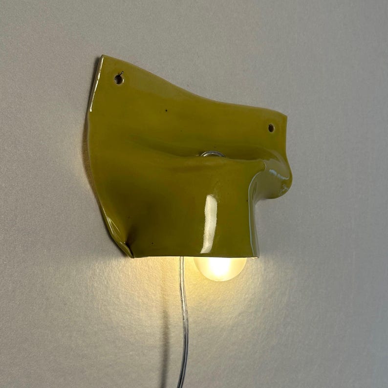 May include: A wall-mounted, olive-green light fixture with a unique, folded design. The fixture has a small, round light bulb at the bottom, and a silver wire extends from the bulb. Two holes are visible at the top for mounting.