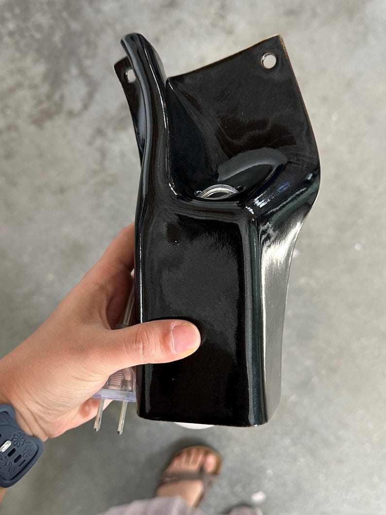May include: A glossy black ceramic wall sconce with a curved, elongated silhouette. The fixture features two circular mounting holes at the top and a central opening designed to hold a light bulb, with a clear electrical cord extending from the base.