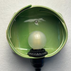 May include: A wall-mounted light fixture featuring a glossy, light green ceramic shade with an organic, folded shape. A black socket holds a round, frosted white light bulb, and a thin, clear electrical cord extends from the bottom base.