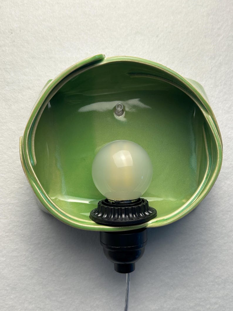 May include: A wall-mounted light fixture featuring a glossy, light green ceramic shade with an organic, folded shape. A black socket holds a round, frosted white light bulb, and a thin, clear electrical cord extends from the bottom base.