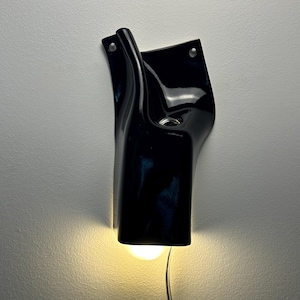 May include: A glossy black ceramic wall sconce with an organic, fluid shape. The fixture features two mounting holes at the top and a downward-facing opening that emits a warm, soft light from an internal bulb, with a thin power cord extending below.