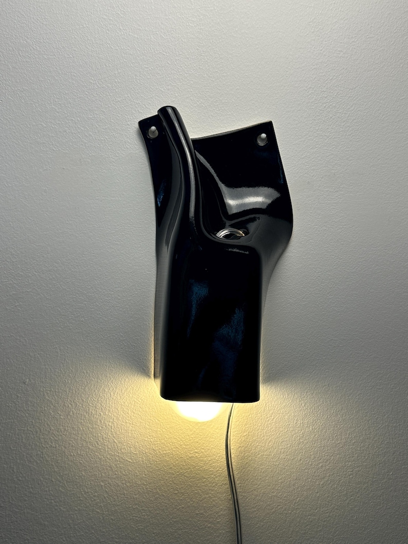 May include: A glossy black ceramic wall sconce with an organic, fluid shape. The fixture features two mounting holes at the top and a downward-facing opening that emits a warm, soft light from an internal bulb, with a thin power cord extending below.