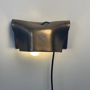 Pode incluir: Um candeeiro de parede com um abajur curvo em tom de bronze met&aacute;lico com acabamento texturizado. A lumin&aacute;ria emite um brilho quente e suave de uma l&acirc;mpada posicionada sob o abajur e inclui um cabo de alimenta&ccedil;&atilde;o preto para baixo.